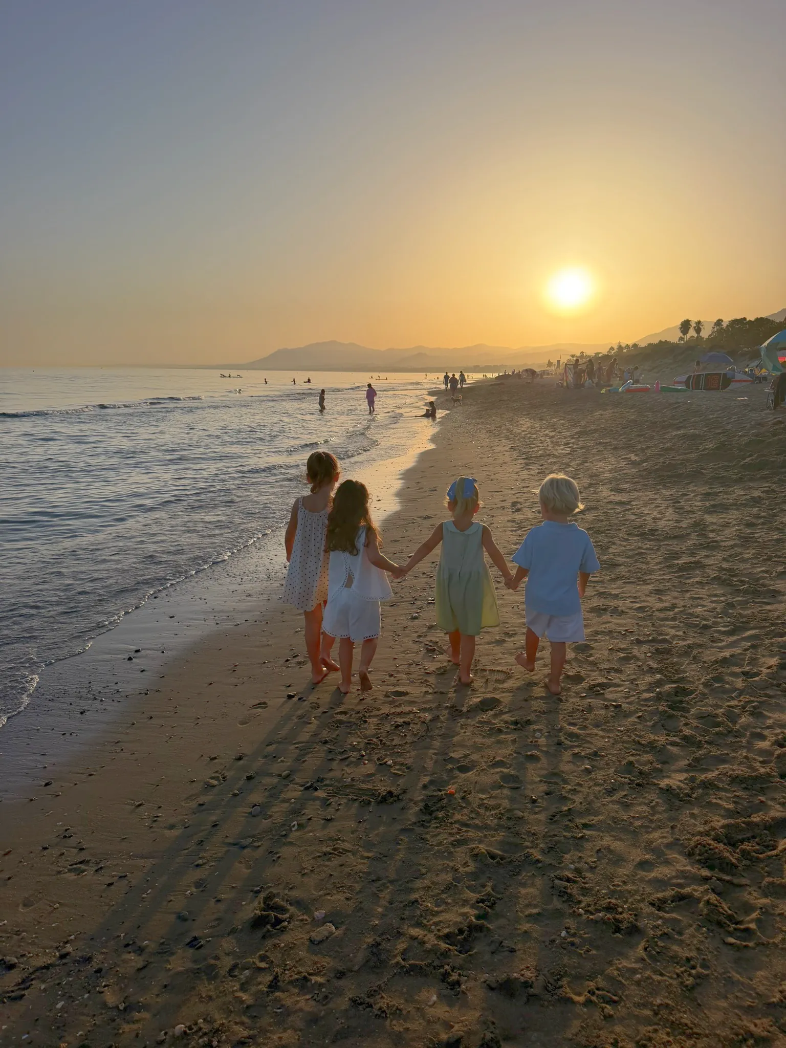 Four children walking hand in hand along the shore at sunset.